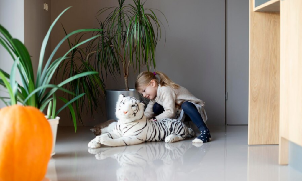 A young girl with blonde hair in a ponytail is kneeling on the floor, hugging a large stuffed white tiger. She is wearing a cozy sweater, dark leggings, and polka-dot socks. The room is decorated with potted plants, and there is a large orange pumpkin in the foreground. The setting appears to be a modern, minimalistic home interior