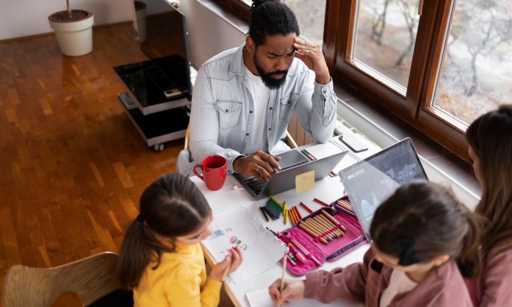 A concerned man working from home, sitting at a desk with a laptop, surrounded by children involved in their activities. The man holds his head in frustration, possibly overwhelmed with multitasking. The desk is filled with notebooks, a red mug, colored pencils, and drawings, illustrating the dynamic of balancing work and family responsibilities