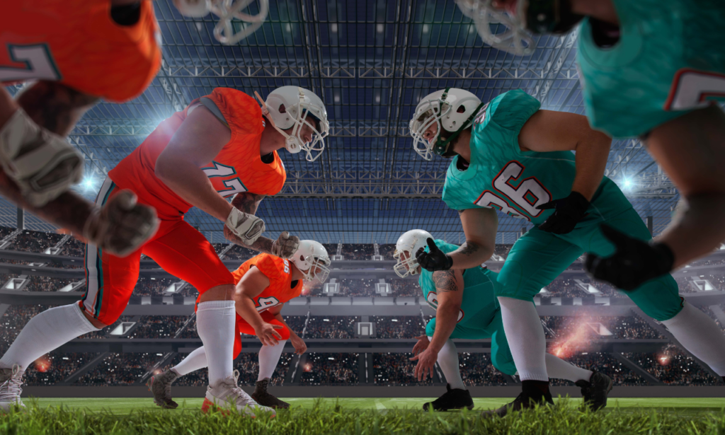 Two American football teams in bright uniforms face off at the line of scrimmage in a packed stadium. One team wears orange jerseys and the other wears teal. The players are in their starting positions, ready for the play, under a large, well-lit stadium dome with a cheering crowd in the background