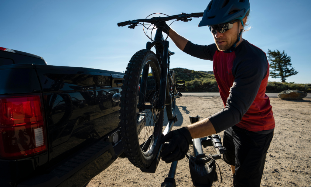 A man wearing a helmet and sunglasses, dressed in cycling gear, is seen loading a mountain bike onto a bike rack attached to the back of a pickup truck. The scene is set outdoors on a sunny day, with clear blue skies and a rugged landscape in the background