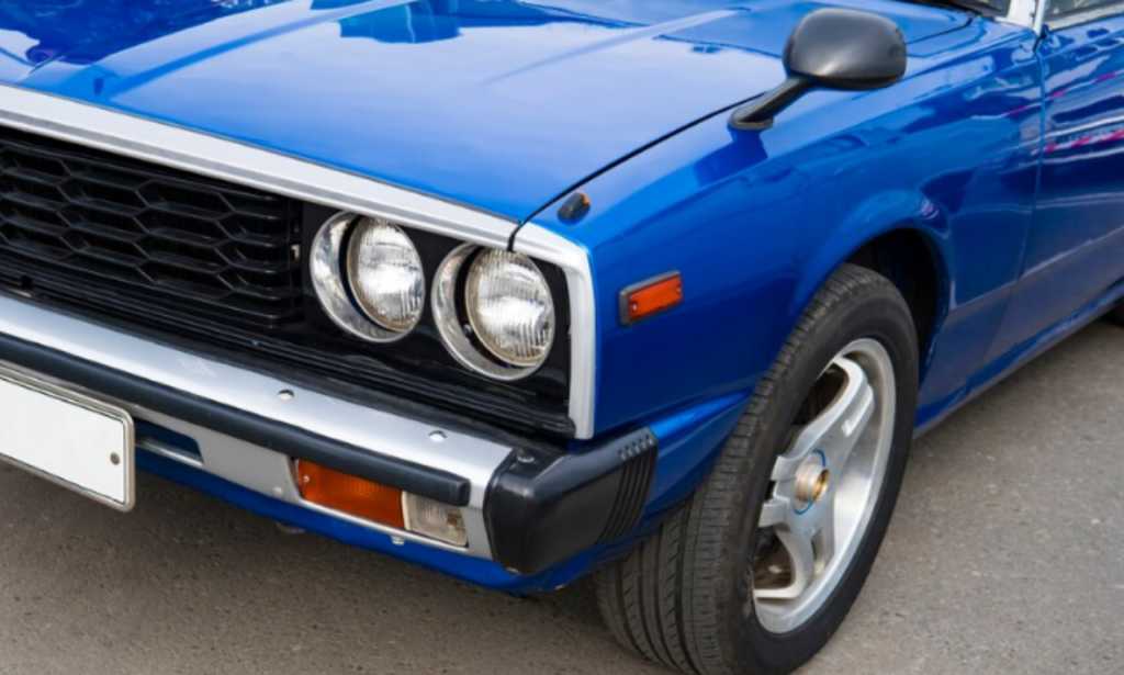 A close-up view of the front end of a classic blue car, showcasing its round headlights, chrome grille, and bumper. The car is parked on a paved surface, emphasizing the vehicle's vintage design and style