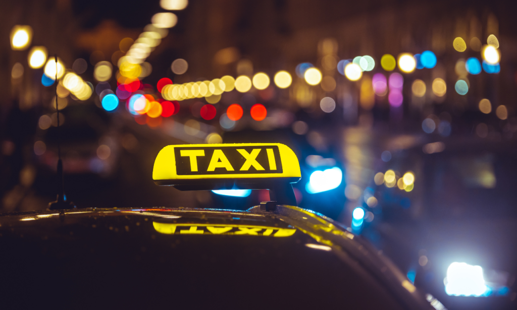 A close-up of a yellow taxi sign illuminated at night, with a bokeh effect of colorful city lights and blurred headlights in the background.