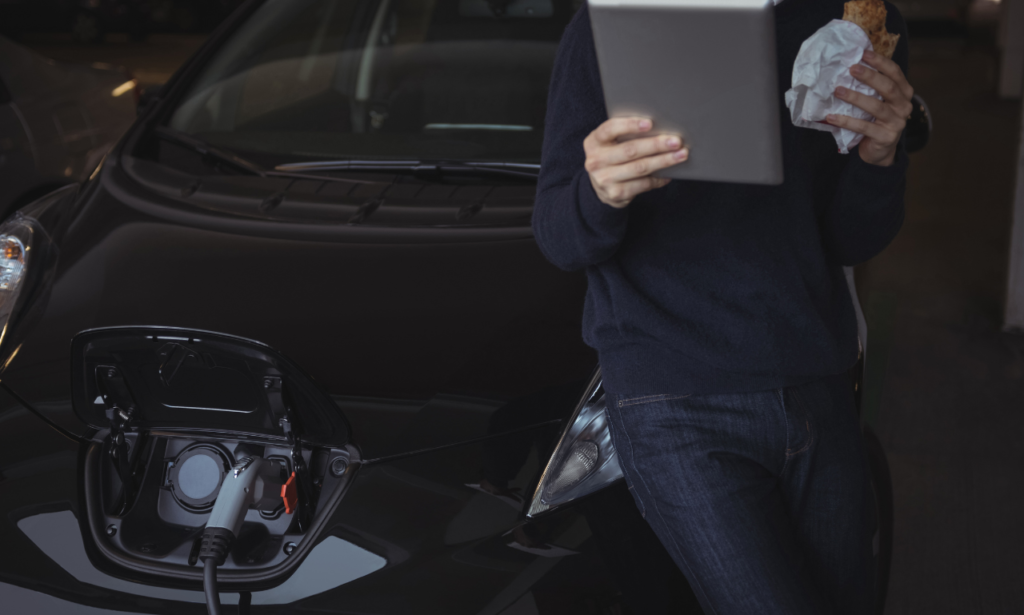 A person leans against an electric car, with a charging port open and a charging cable connected. They hold a tablet in one hand and a wrapped snack in the other, suggesting a casual break while the vehicle charges. The scene captures the modern lifestyle of EV owners, balancing technology, convenience, and eco-consciousness.