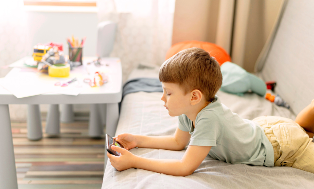 A young boy lying on a bed, engrossed in playing a game on a smartphone. In the background, a small table holds colorful stationery, toys, and art supplies, creating a casual and creative environment.