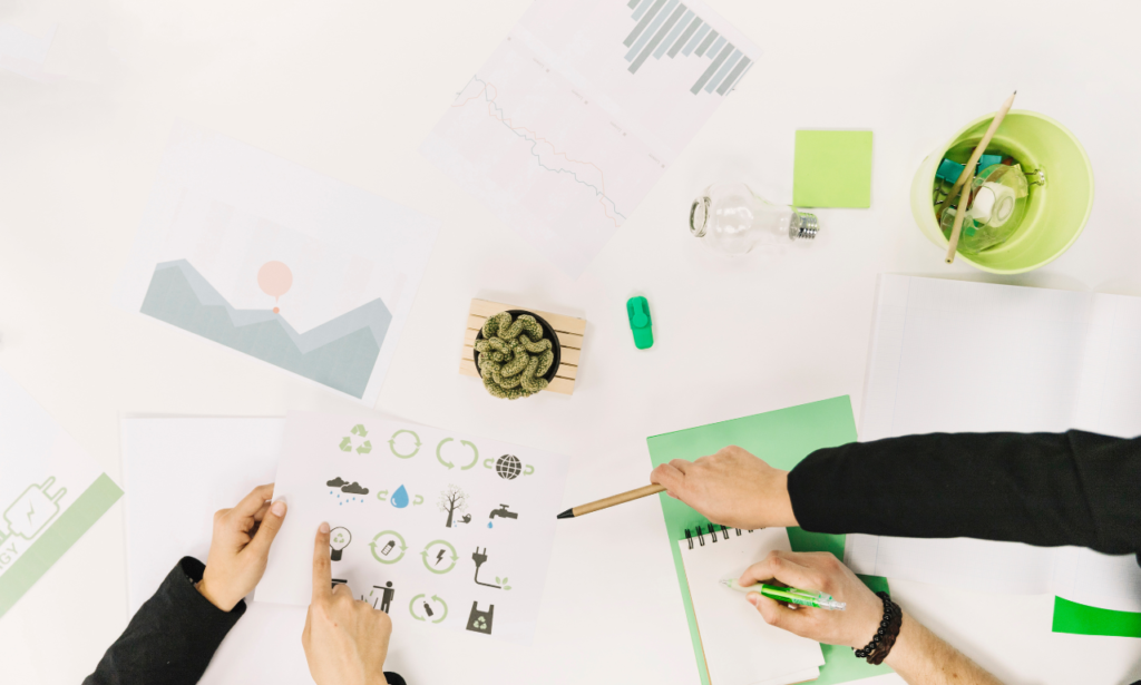 A workspace featuring hands collaborating over eco-friendly-themed documents, including icons of sustainability, recycling, and renewable energy. The desk is adorned with a potted cactus, green stationery, and charts, symbolizing environmental consciousness and teamwork.