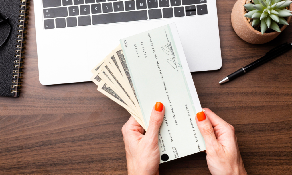 Close-up image of hands holding a stack of dollar bills and a check in front of a laptop. The check is from a rebate program, showcasing a business or financial transaction setting. The workspace includes a notepad, pen, and a small potted succulent plant, creating a professional yet organized atmosphere