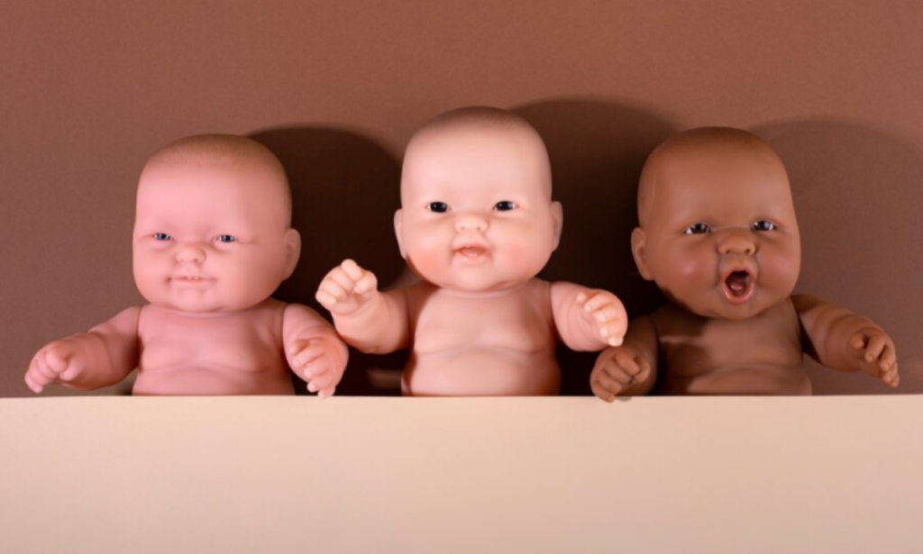 Three lifelike silicone baby dolls in various skin tones arranged in a row against a warm, neutral background. Each doll has distinct facial expressions, showcasing realistic details and textures that enhance their lifelike appearance.